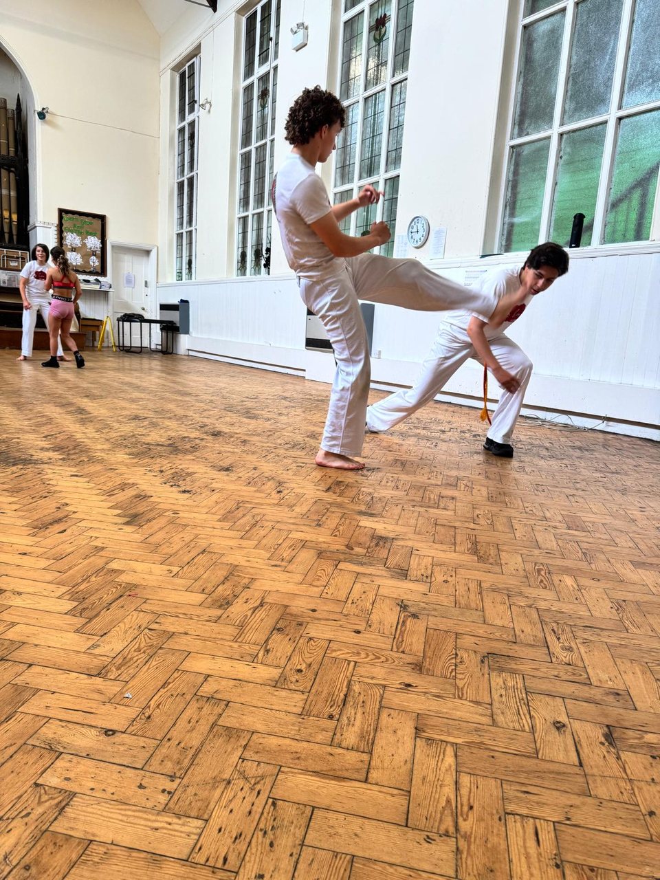 Two young capoeiristas practise a kick and an escape in a bright hall with a wooden floor.